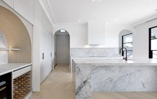 Architectural Joinery | Bespoke Wine Cellar High-end kitchen island and bespoke wine cellar featuring waterfall benchtop in Super White Dolomite, custom timber dowels, and architectural arch with a Coastal Oak Woodmatt finish.